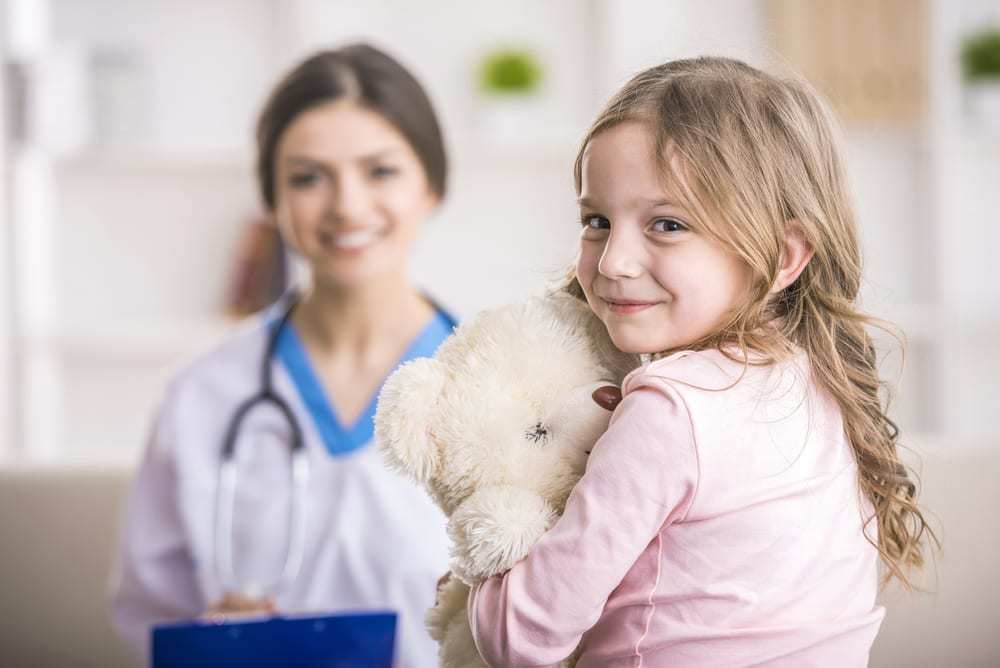 Little girl with teddy bear smiling at camera, doctor in background