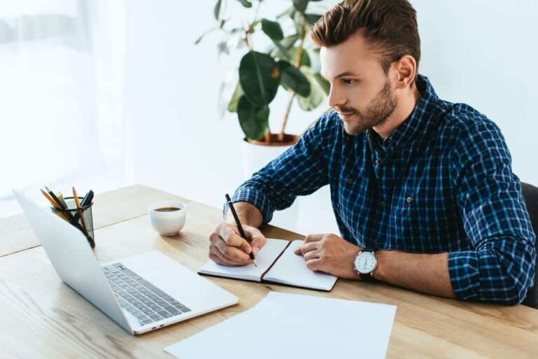 Young man taking notes while looking at laptop