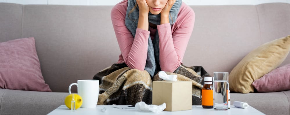 Young woman on couch, ill, with tissues, medicine, water, and tea in front of her