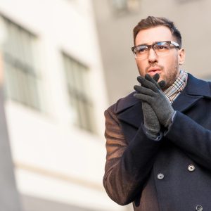 Young man dressed smartly, outside in cold weather, blowing on hands