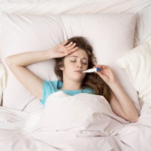 Young woman sick with the flu, in bed with thermometer